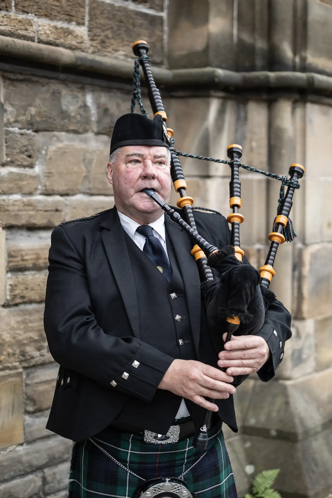 A man in traditional scottish attire playing the bagpipes outdoors, featuring a tartan kilt and a balmoral bonnet.