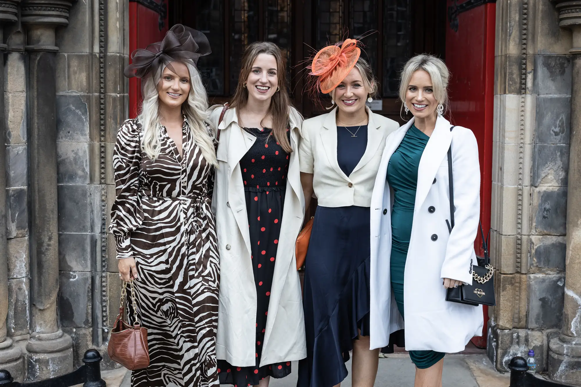 Four women in stylish outfits and colorful hats smiling outside a historic building.