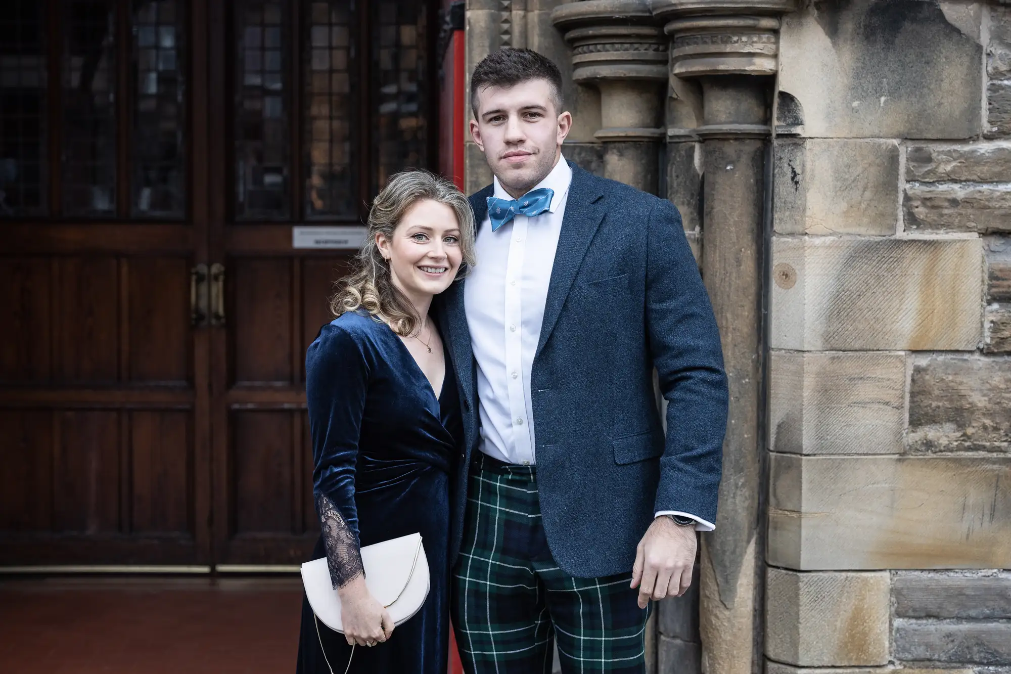 A couple posing in formal attire, the woman in a blue velvet dress and the man in a kilt and blazer, in front of a historic building's wooden doors.