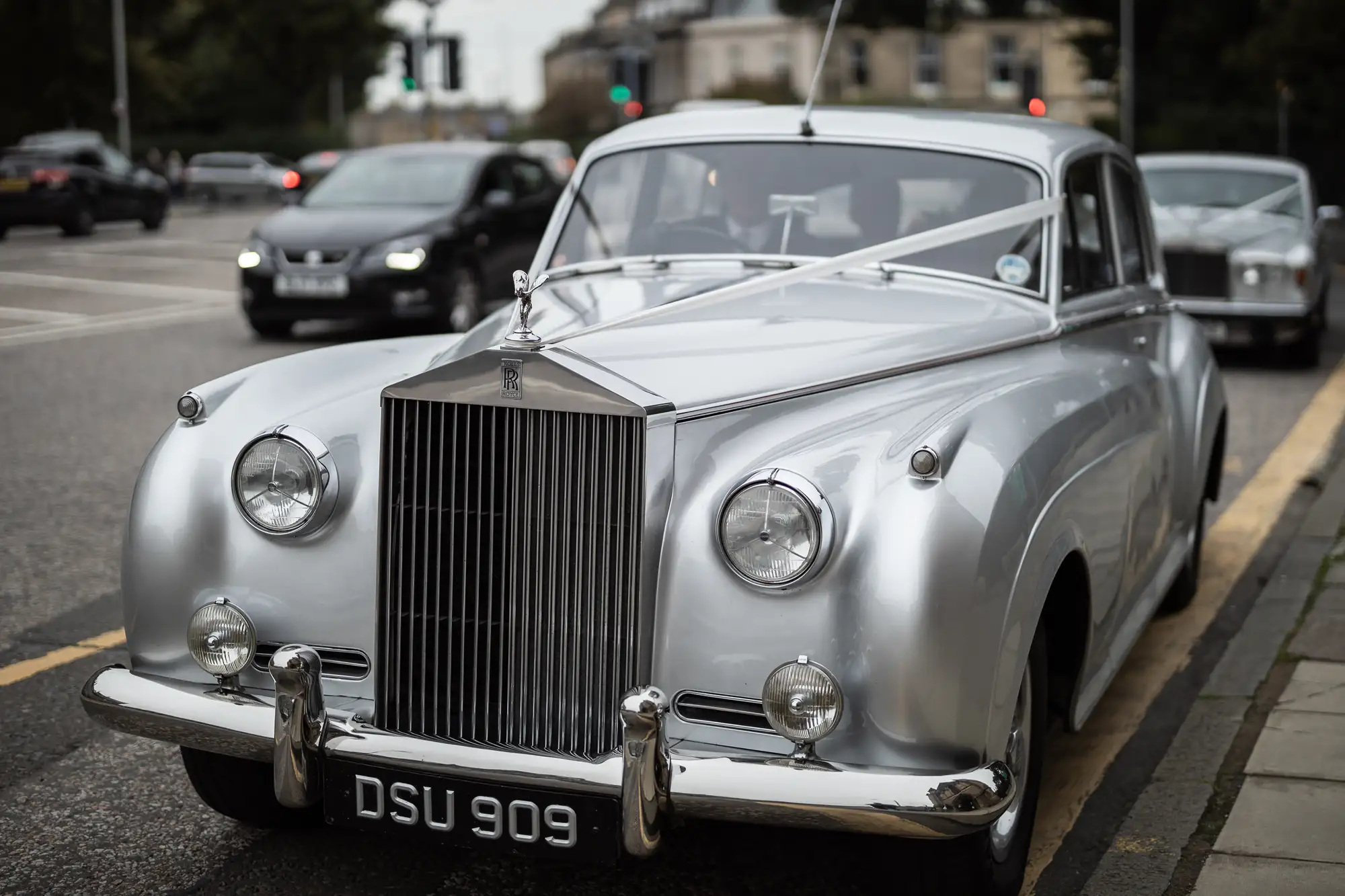 A vintage silver rolls-royce parked on a city street, with modern cars in the background.