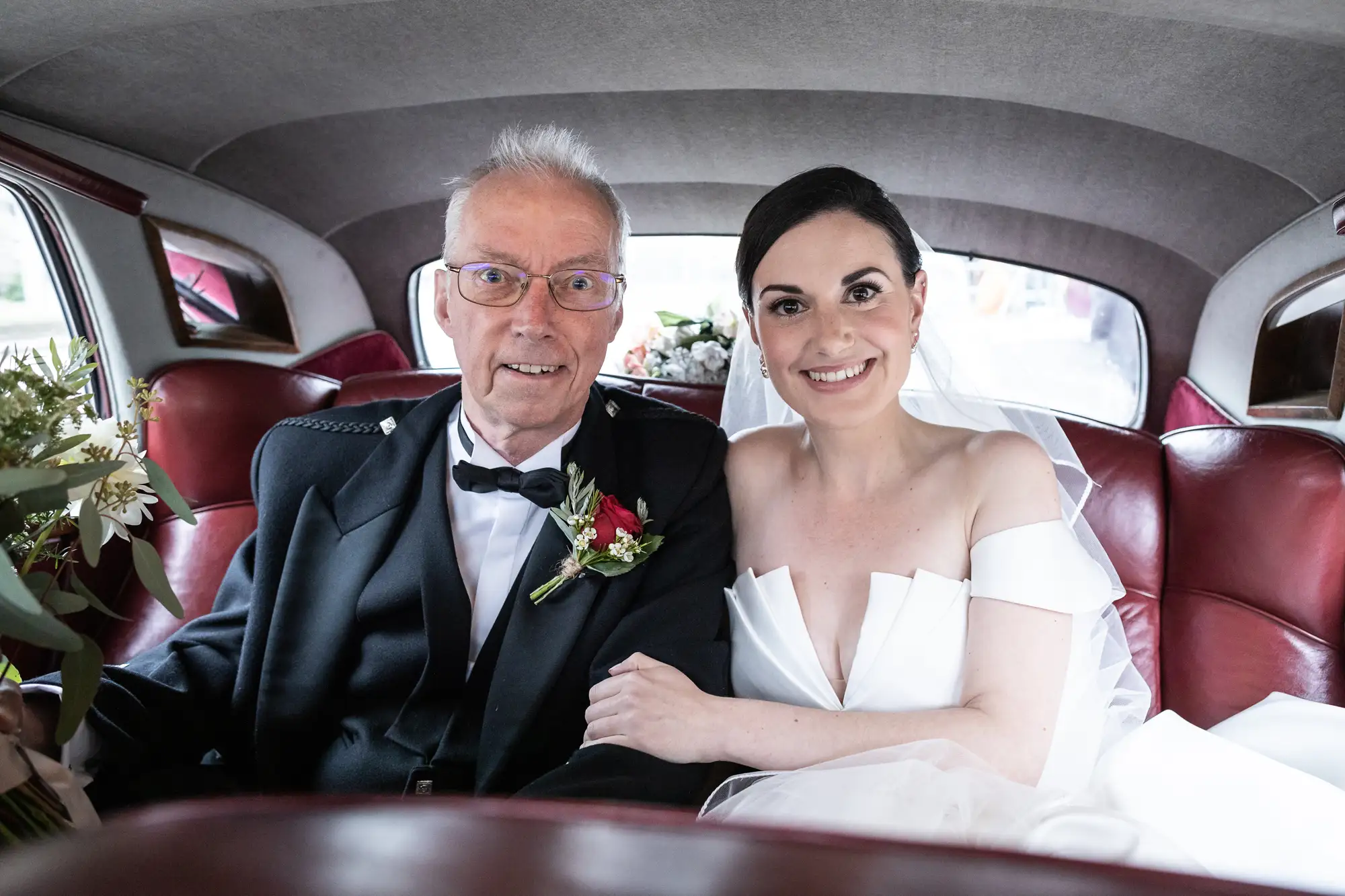 An elderly man and a young woman in formal attire smiling inside a vintage car with red leather seats.