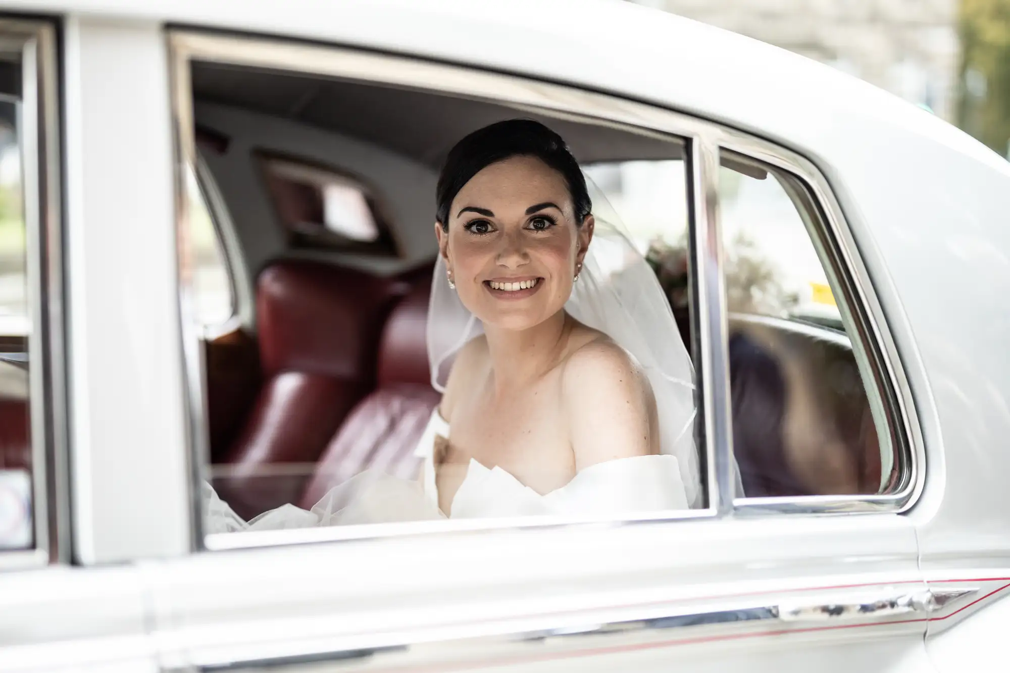 A bride smiling from the backseat of a vintage car with a white exterior and red leather interior.