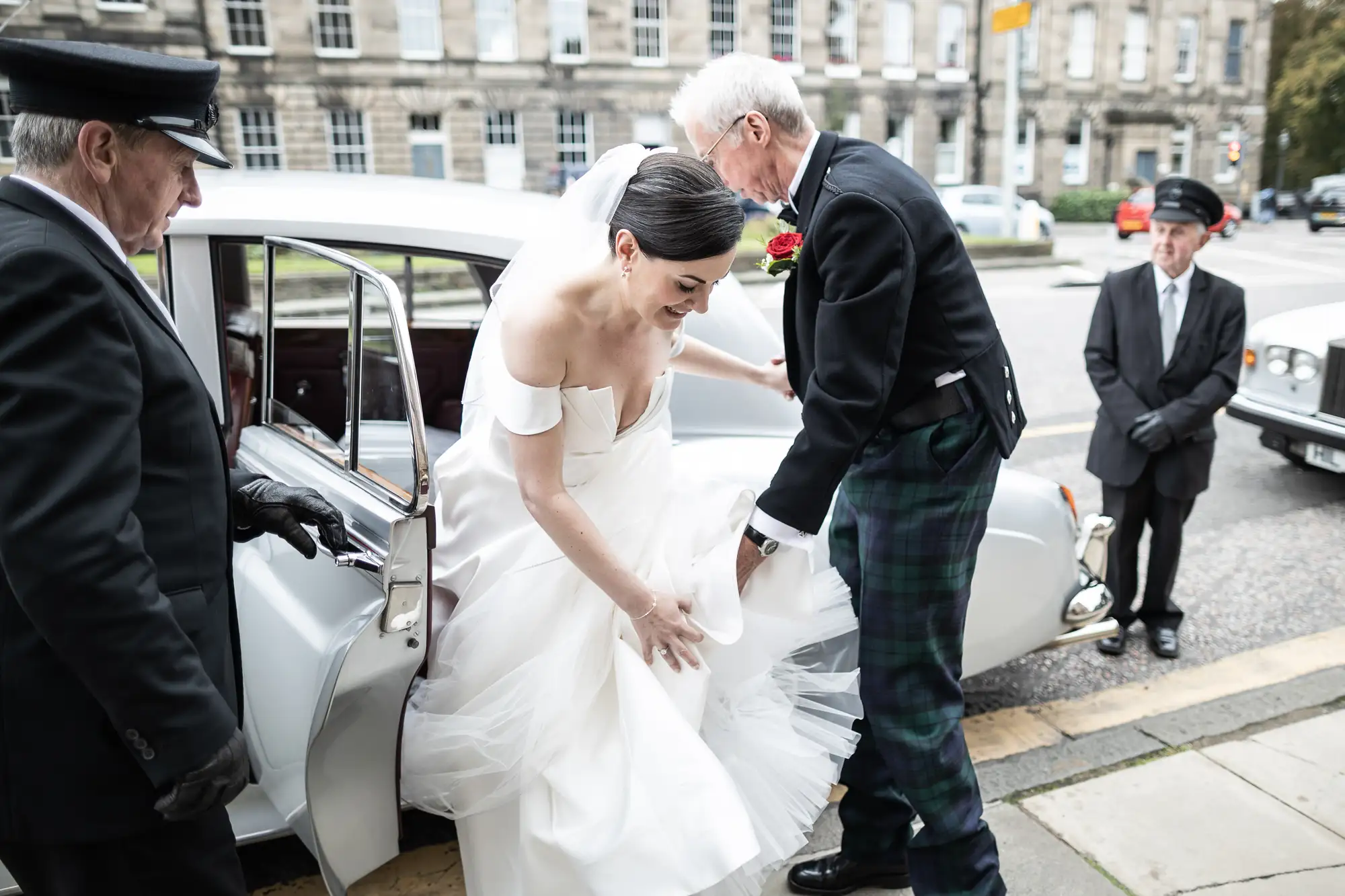 A bride in a white dress exiting a vintage car with assistance from an elderly man, while two men in formal attire observe.
