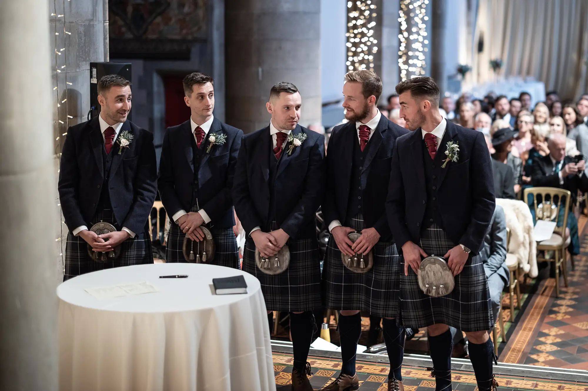 Five groomsmen in kilts standing in a church during a wedding ceremony, with guests seated in the background.