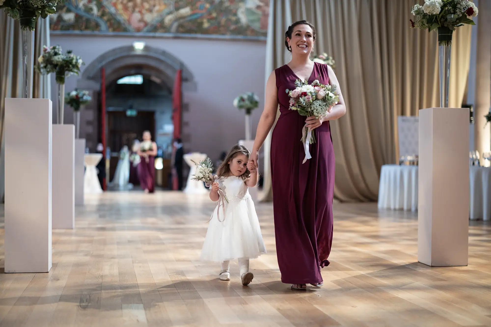 A woman in a long purple dress smiles as she walks down an aisle with a young girl in a white dress, both holding bouquets, in a decorated hall.