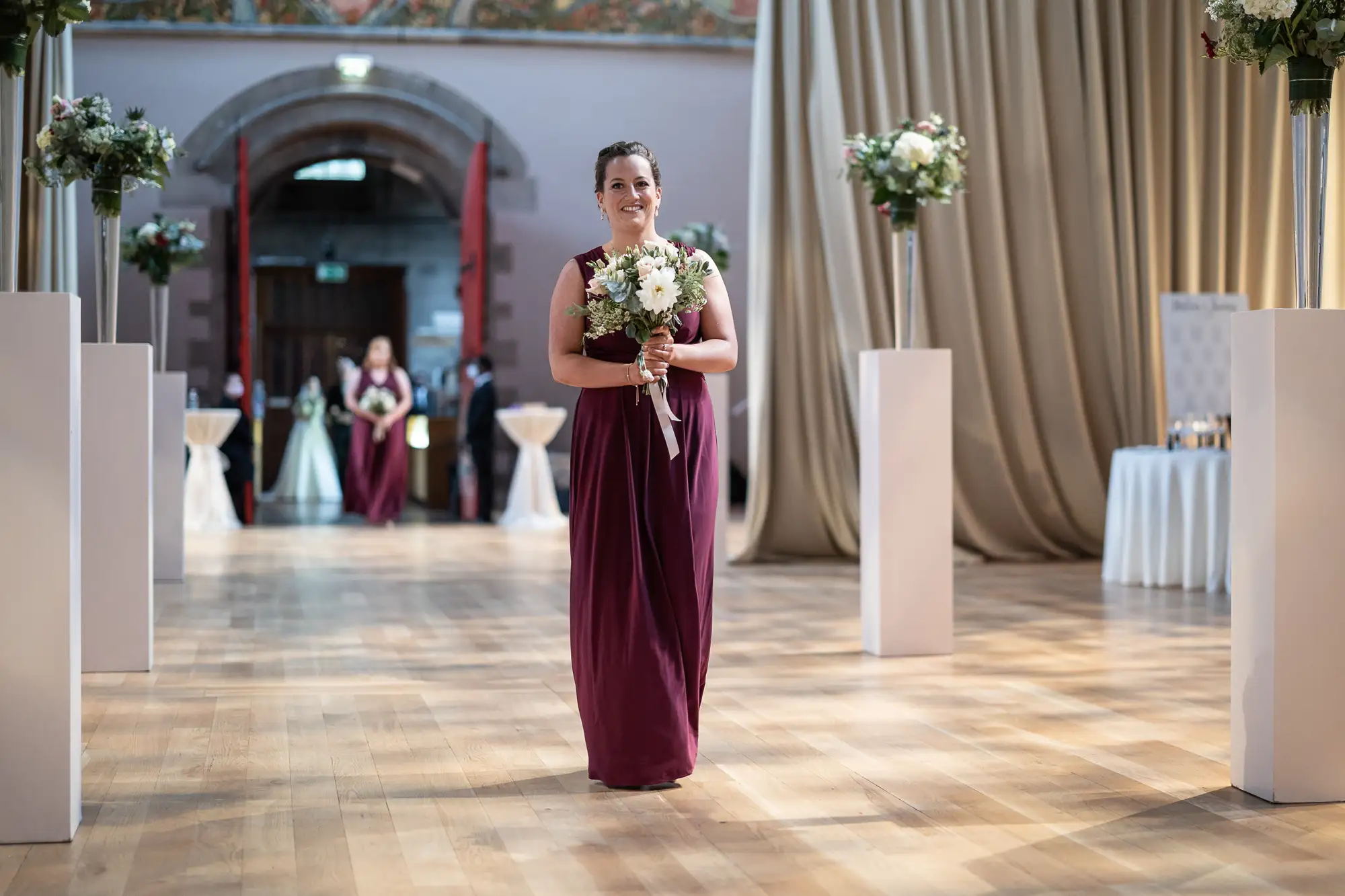 A woman in a burgundy dress smiling and holding a bouquet at a wedding ceremony in an elegant hall.