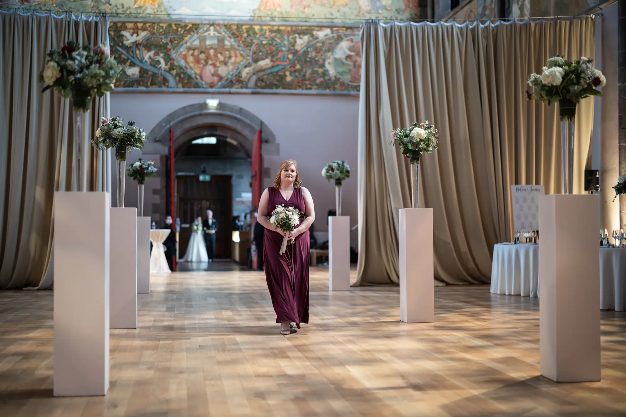 A woman in a burgundy dress holding a bouquet walks between rows of floral arrangements in an ornately decorated hall.