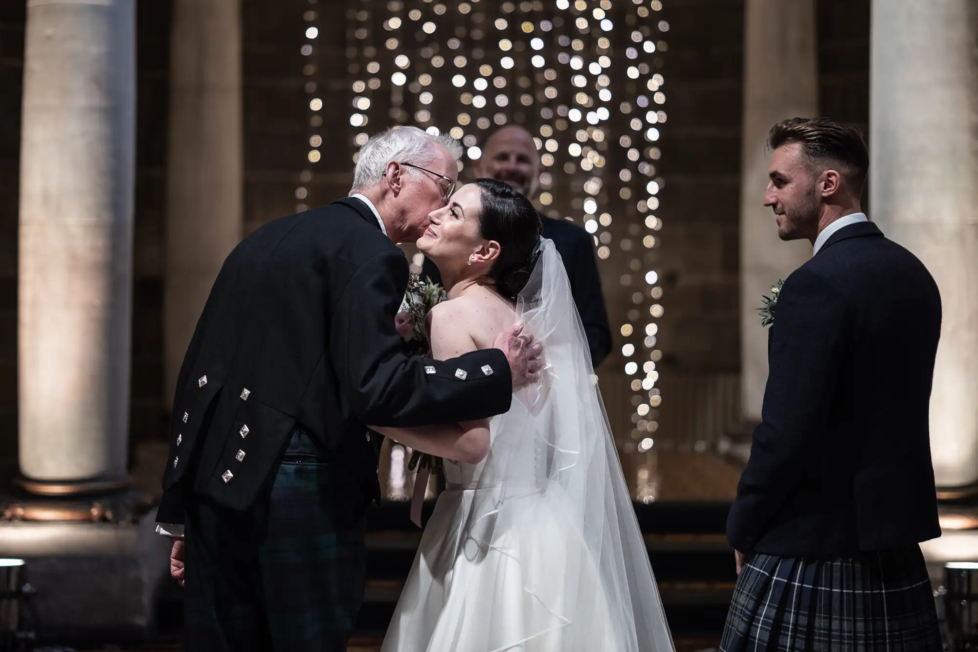 An elderly man in a kilt kisses the cheek of a bride in a white dress, while a groom in a kilt watches, all against a background of twinkling lights in a grand hall.