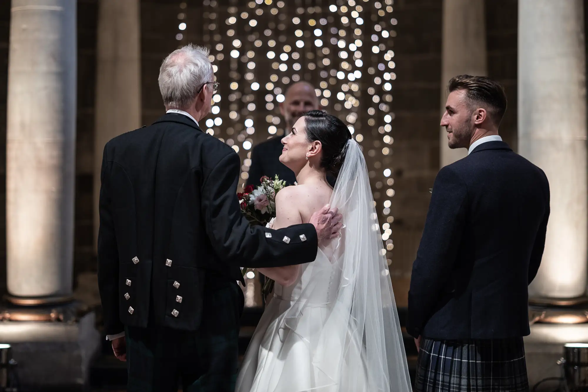 A bride in a white dress and a groom in a kilt stand at the altar, exchanging vows in front of an officiant, with twinkling lights in the background.