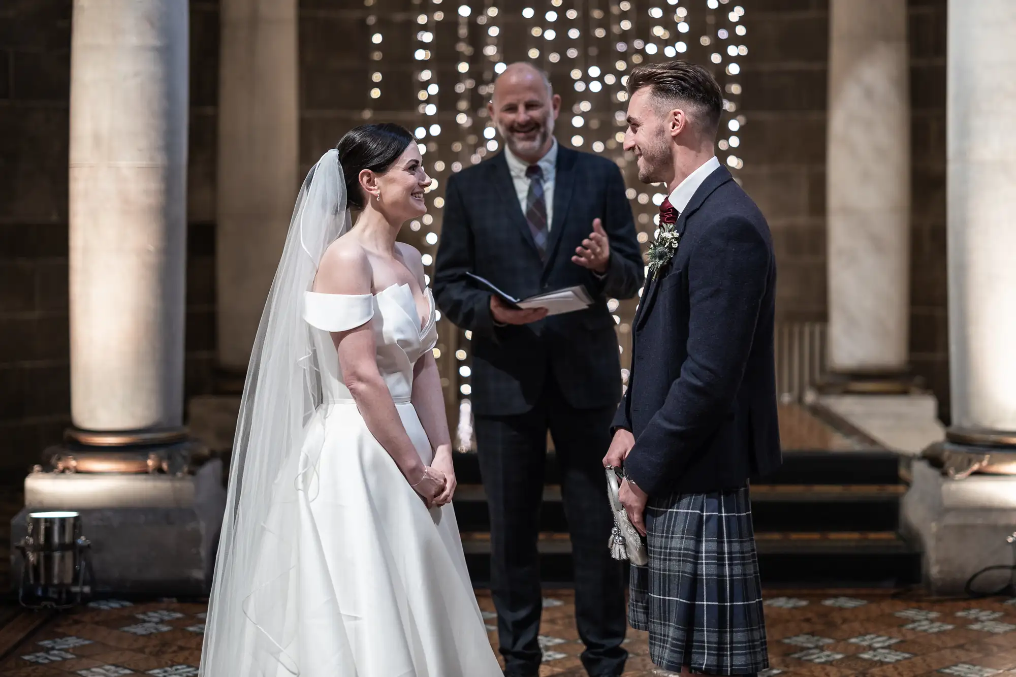 A bride in a white gown and a groom in a kilt exchanging vows in a church, with an officiant smiling behind them and fairy lights in the background.