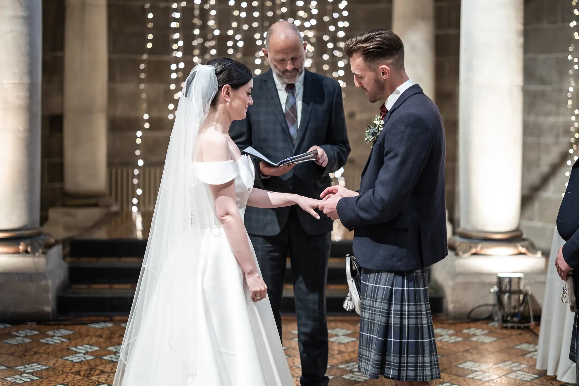 A bride in a white dress and a groom in a kilt hold hands during their wedding ceremony, with an officiant reading from a book in a church lit by string lights.