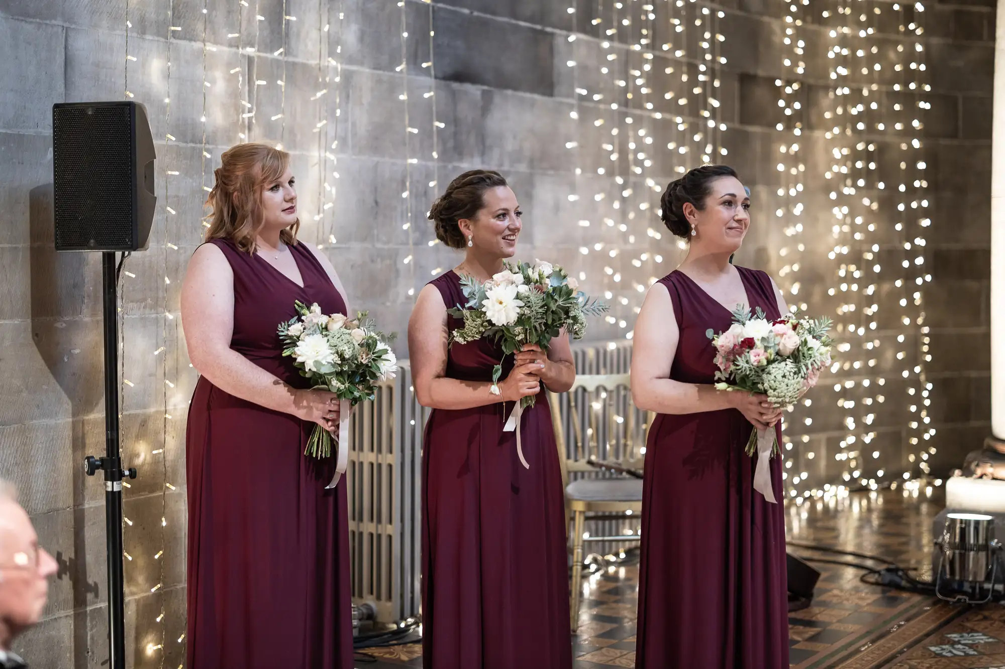Three bridesmaids in burgundy dresses holding bouquets, standing in front of a curtain of fairy lights at a wedding.
