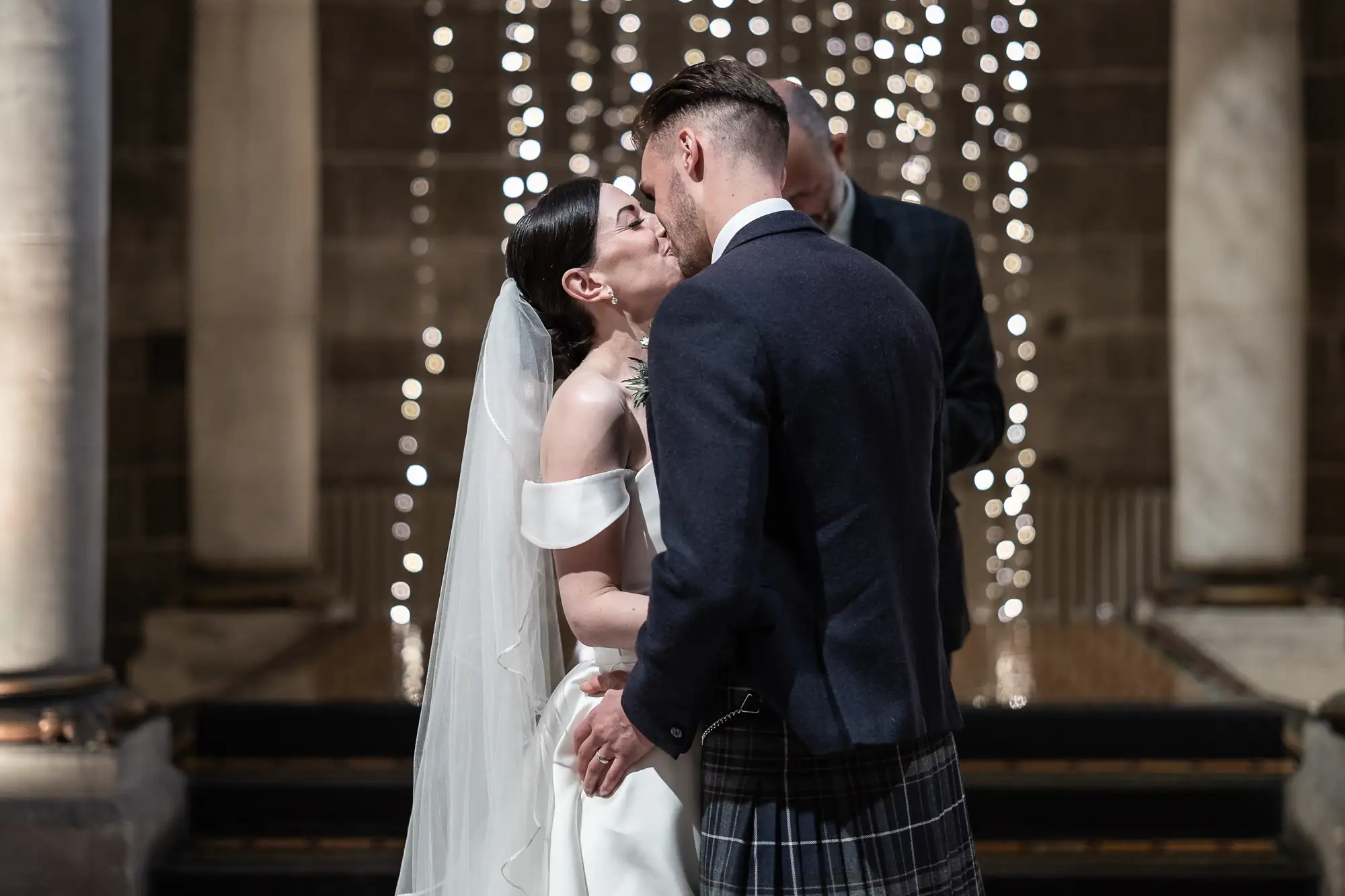 Bride and groom kissing in a church adorned with string lights, the bride in a white dress and veil, and the groom in a kilt and jacket.