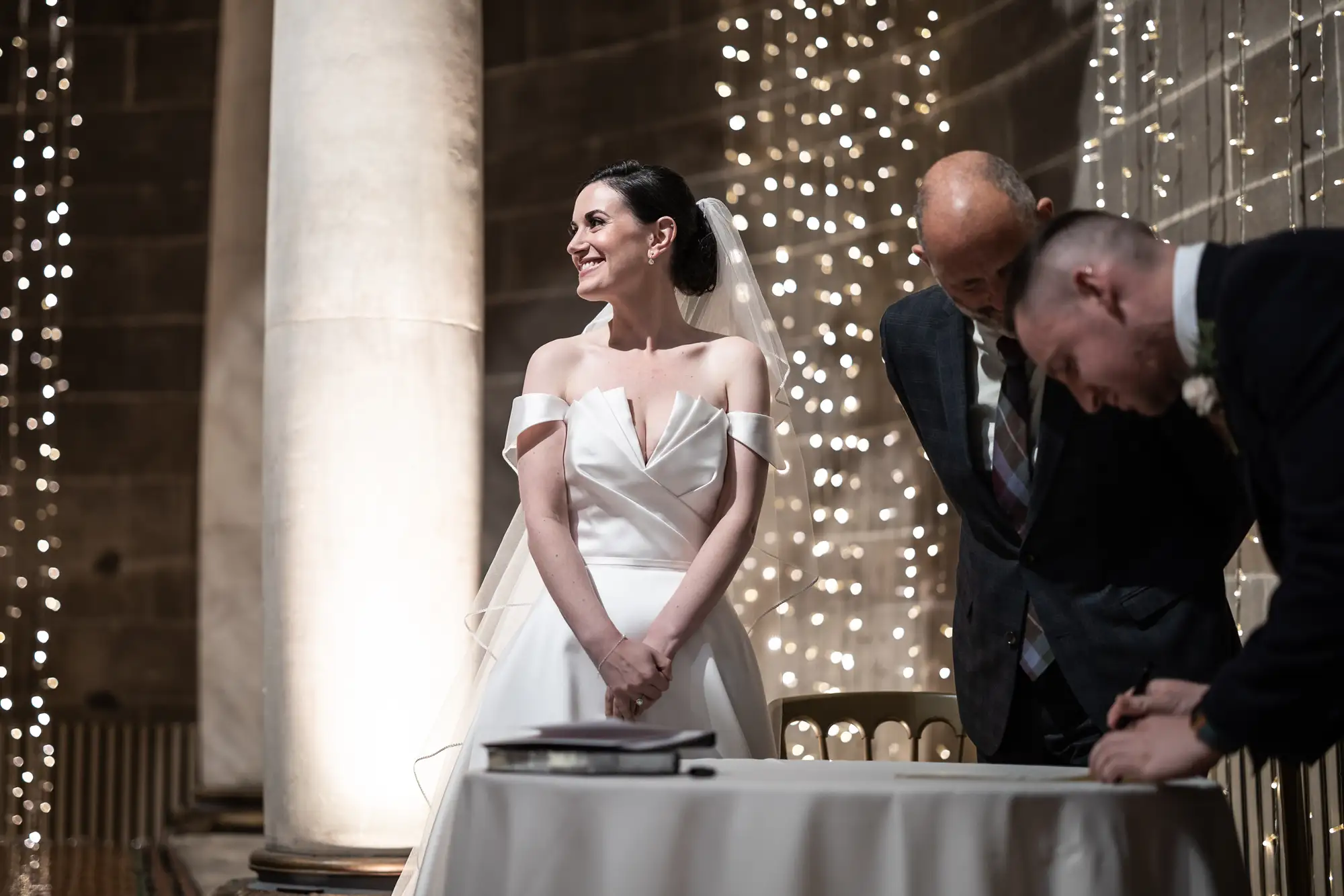 A bride in a white off-the-shoulder gown smiles during a ceremony, standing near two men signing a document, with a background of twinkling lights.