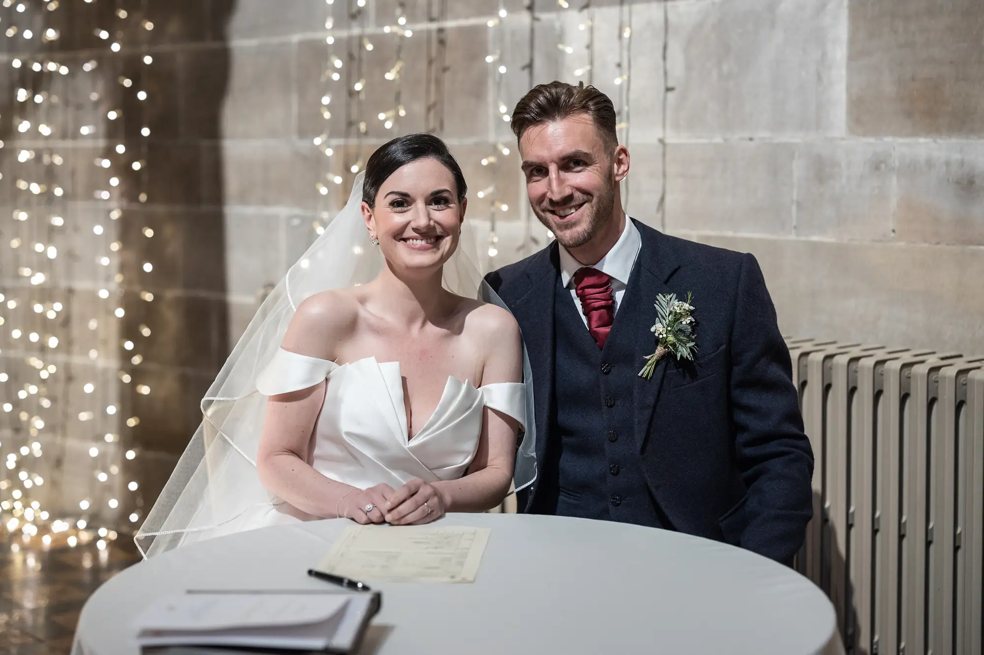 A bride in a white dress and a groom in a dark suit smile happily at a signing table, with a backdrop of twinkling lights.