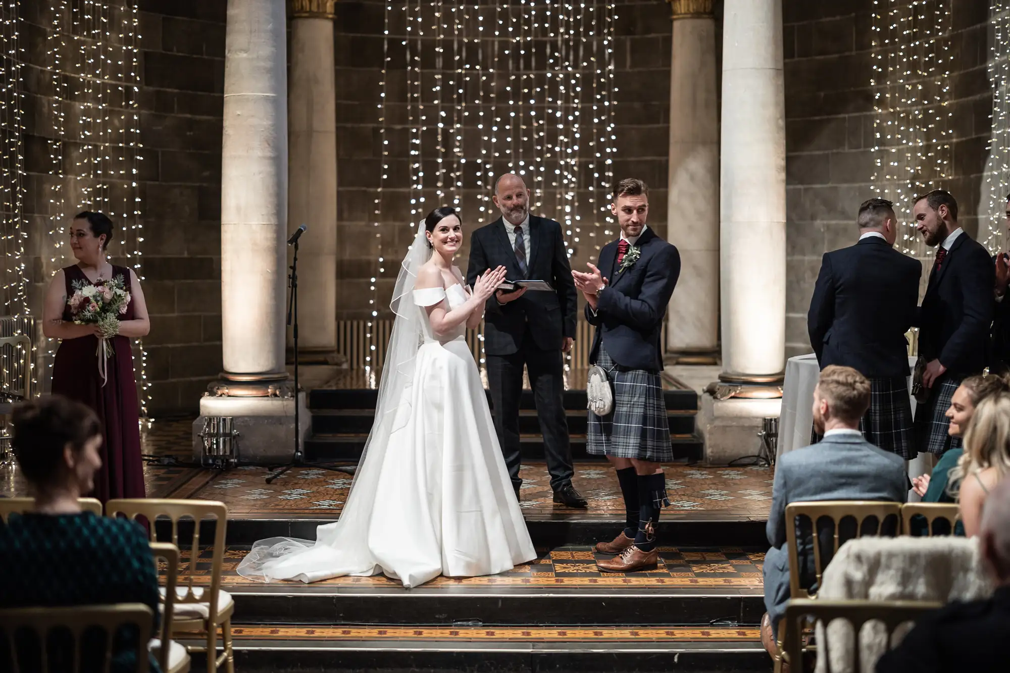Bride and groom smiling at their wedding ceremony in a grand hall decorated with fairy lights, surrounded by guests and a man in a kilt clapping.