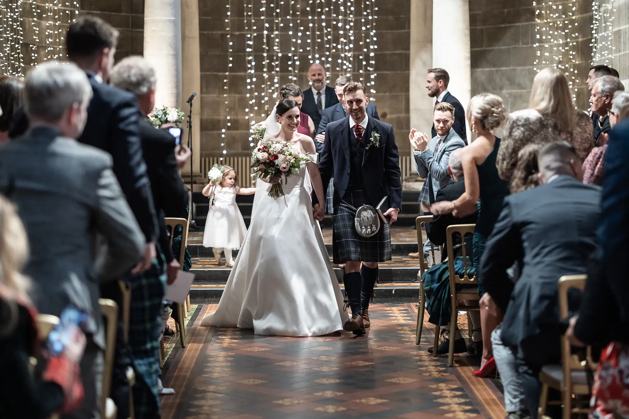 A bride and groom walking down the aisle in a church, the groom in a kilt, surrounded by guests, with string lights and candles in the background.