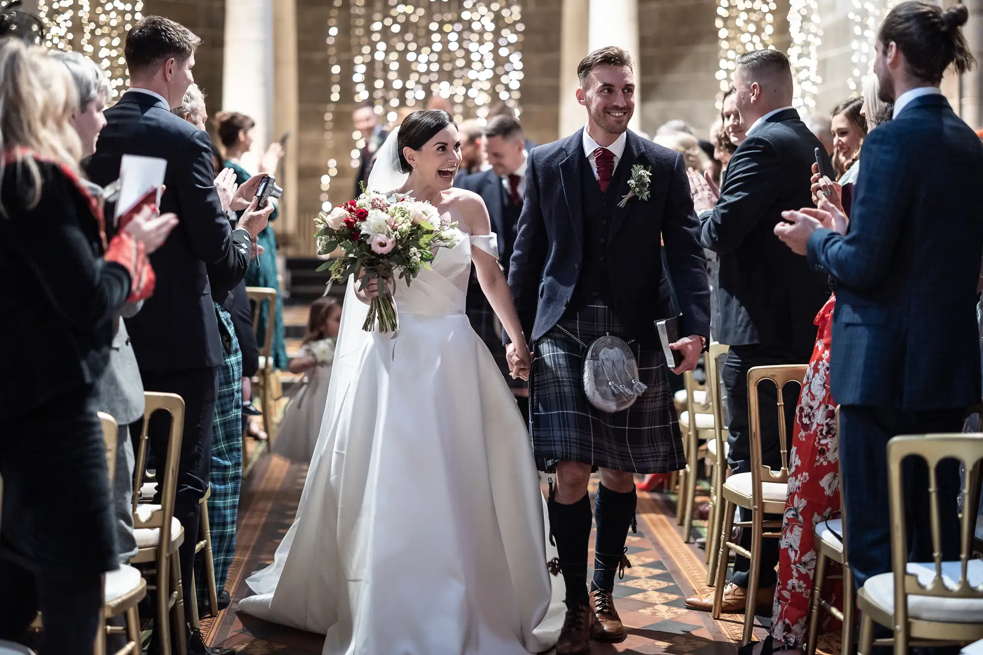 A bride and groom smiling as they walk through a wedding aisle, surrounded by applauding guests, the groom wearing a kilt and the bride holding a bouquet.