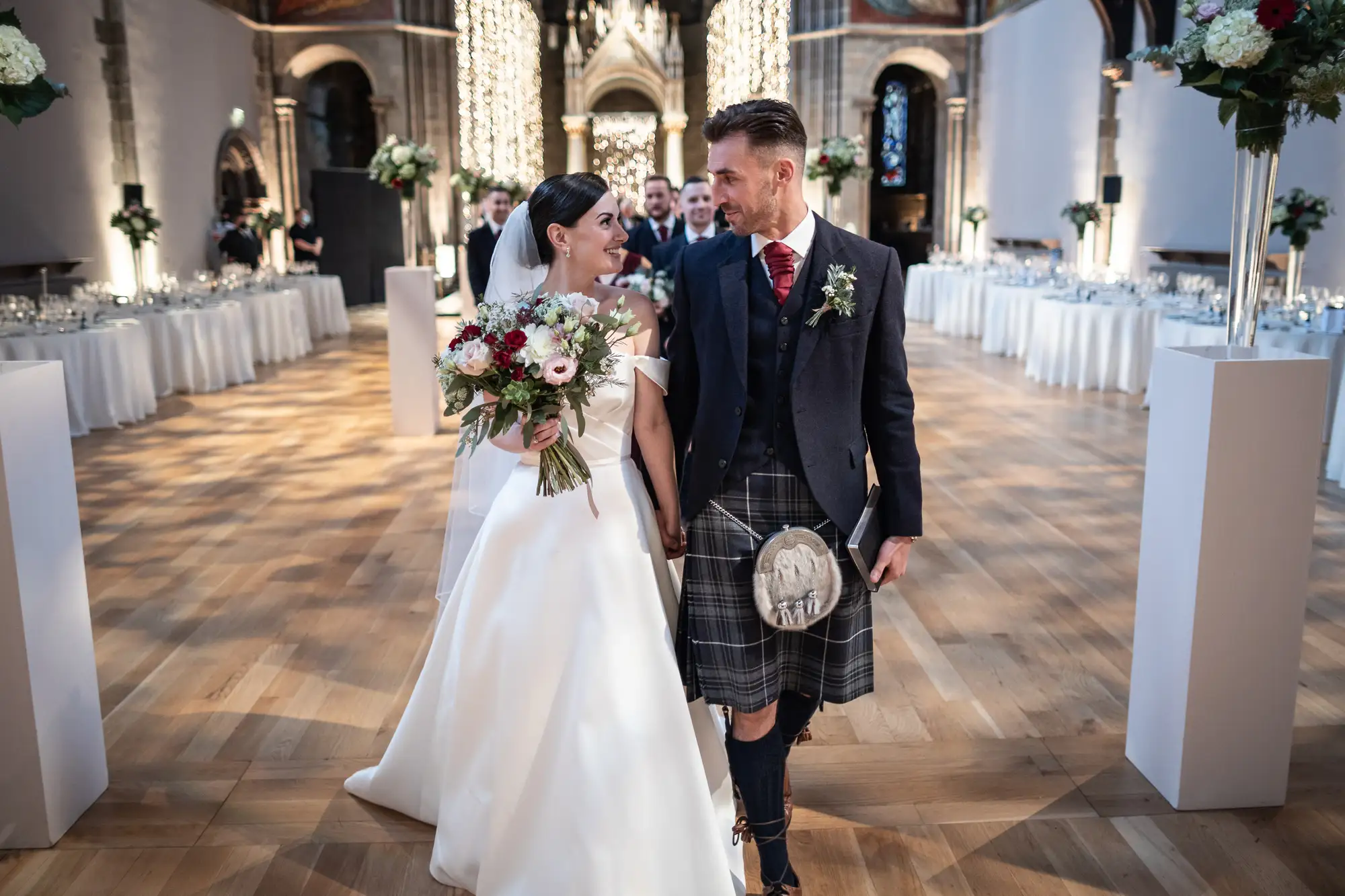 A bride in a white dress and a groom in a kilt walk hand in hand through a wedding venue, decorated with white columns and banquet tables.