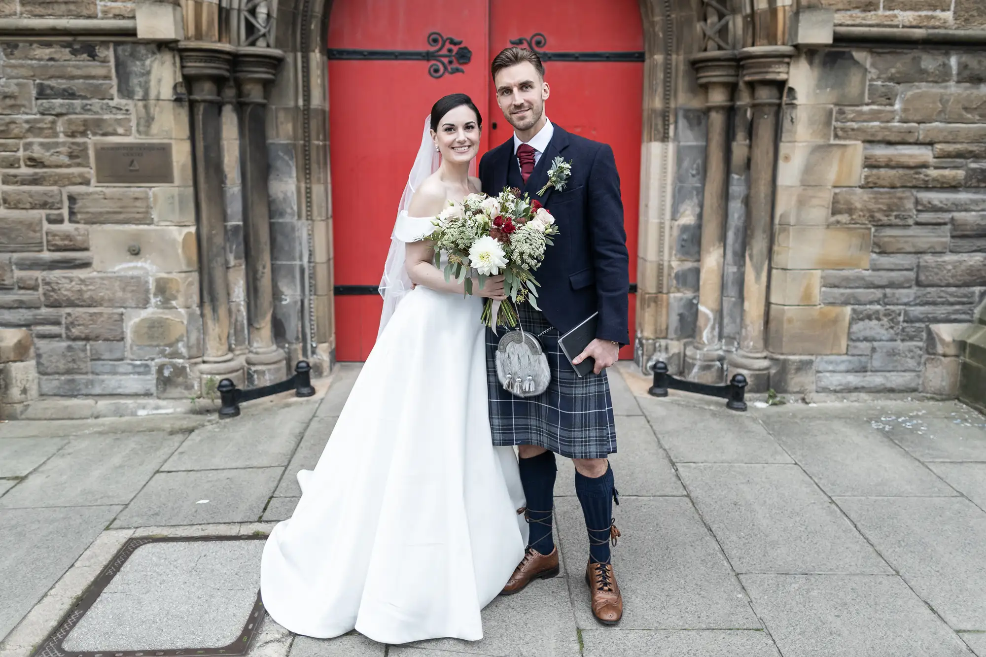 A bride in a white gown and a groom in a kilt holding a bouquet, standing in front of a red door at a stone church.