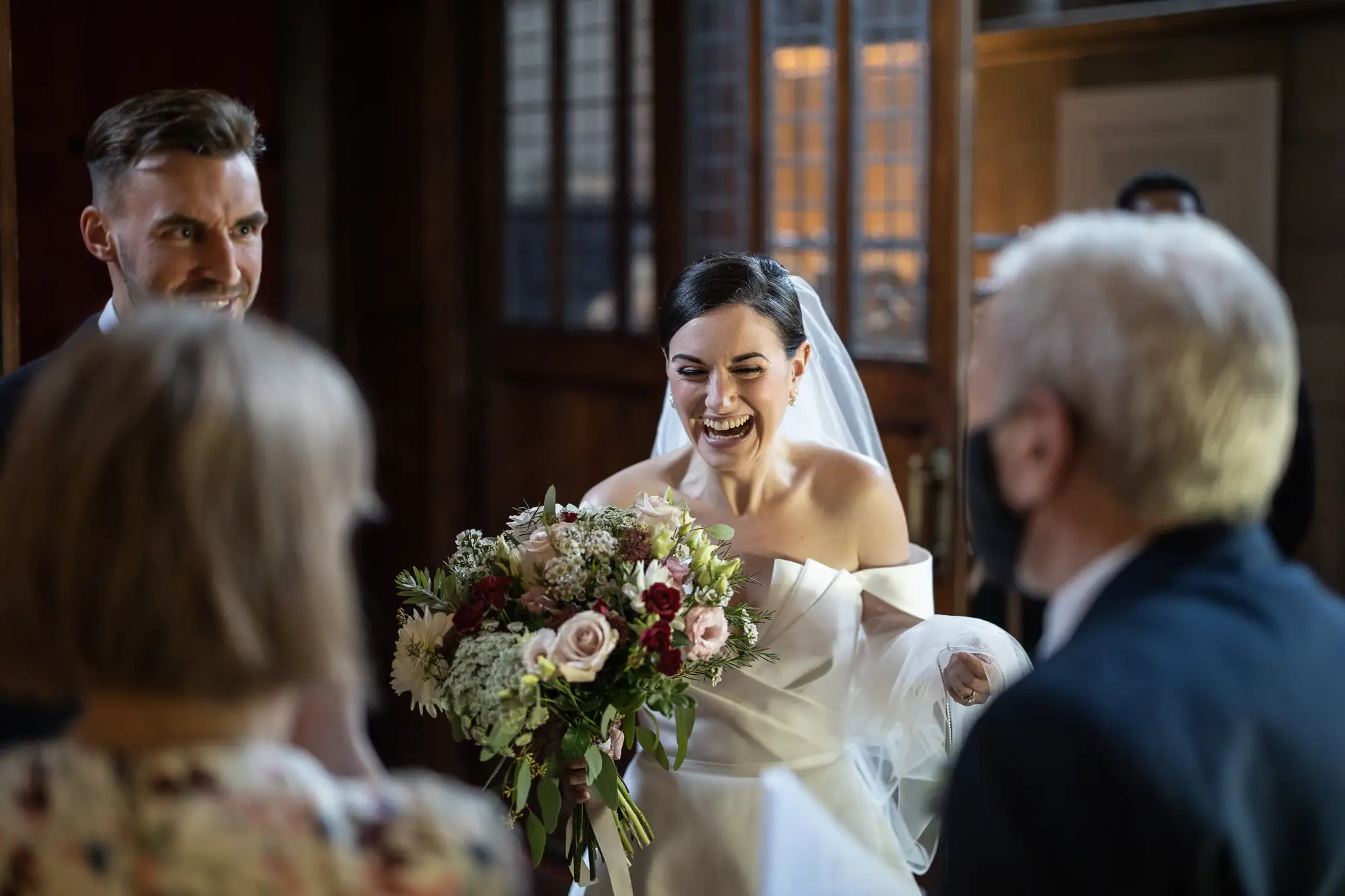 A bride laughing joyfully, holding a bouquet, with a groom and guests around her during a wedding ceremony indoors.