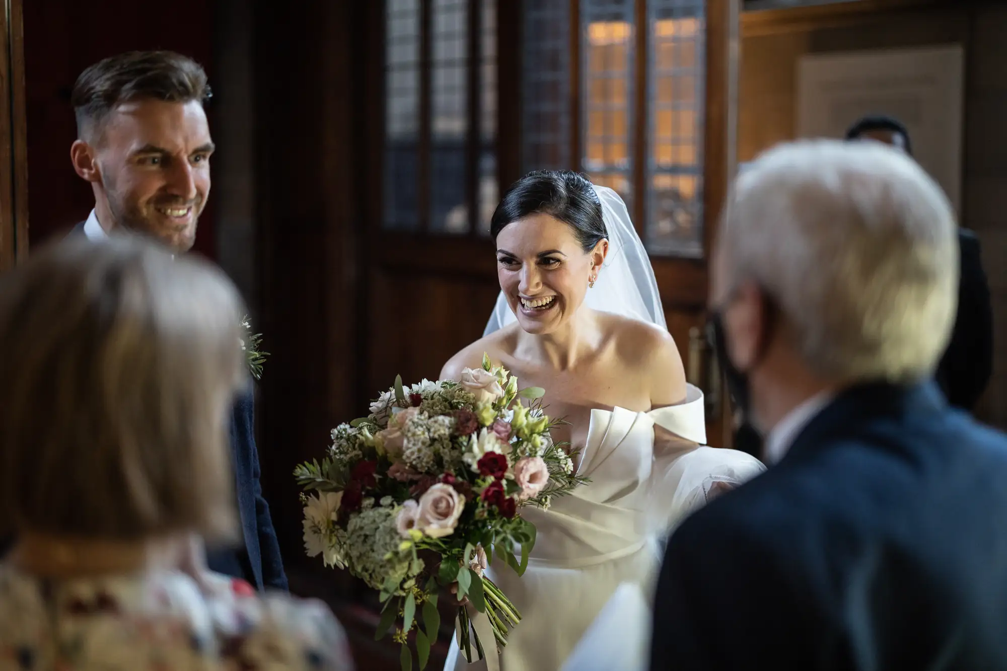 A bride and groom smiling joyously, surrounded by guests in a warmly lit indoor setting. the bride holds a bouquet of flowers.