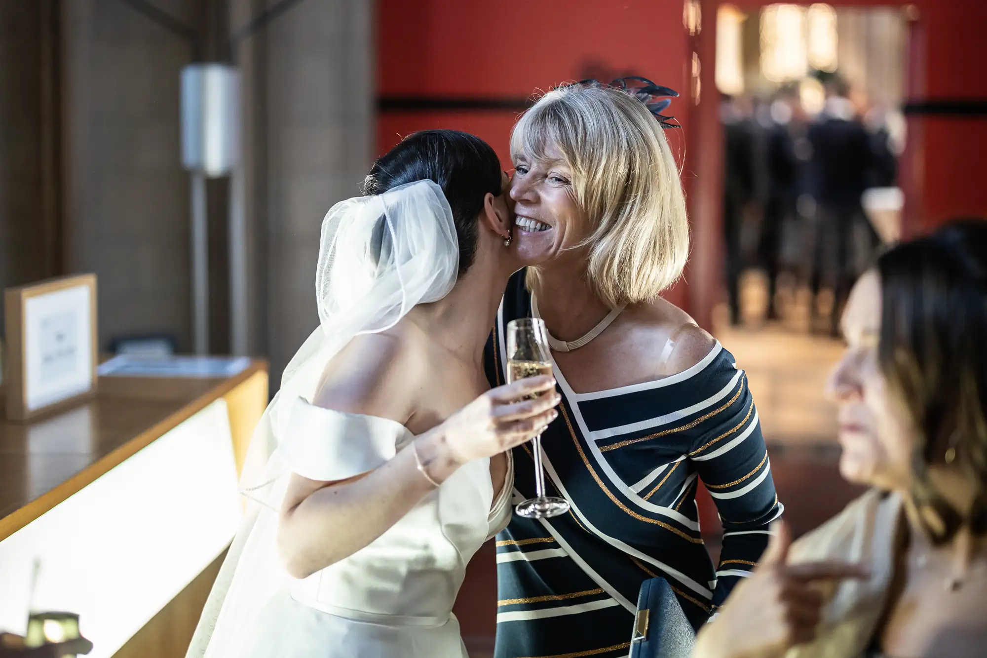 A bride in a white off-shoulder dress embraces an older woman holding a champagne glass at a wedding reception.