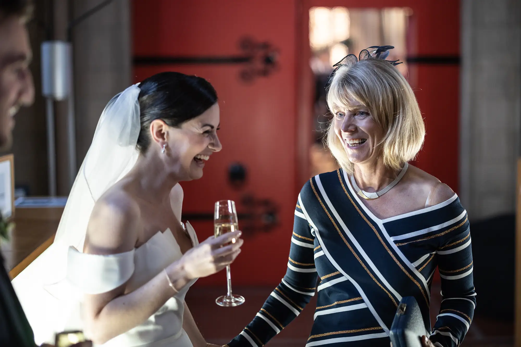 A bride and an older woman smiling and talking indoors at a wedding, both holding glasses of champagne.