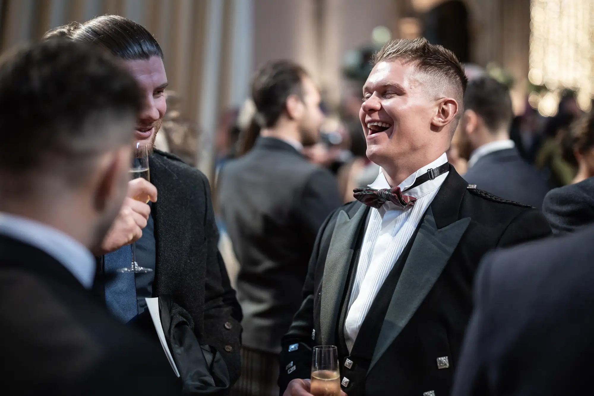 Two men in formal attire laugh and chat at a social event, one holding a glass of champagne.