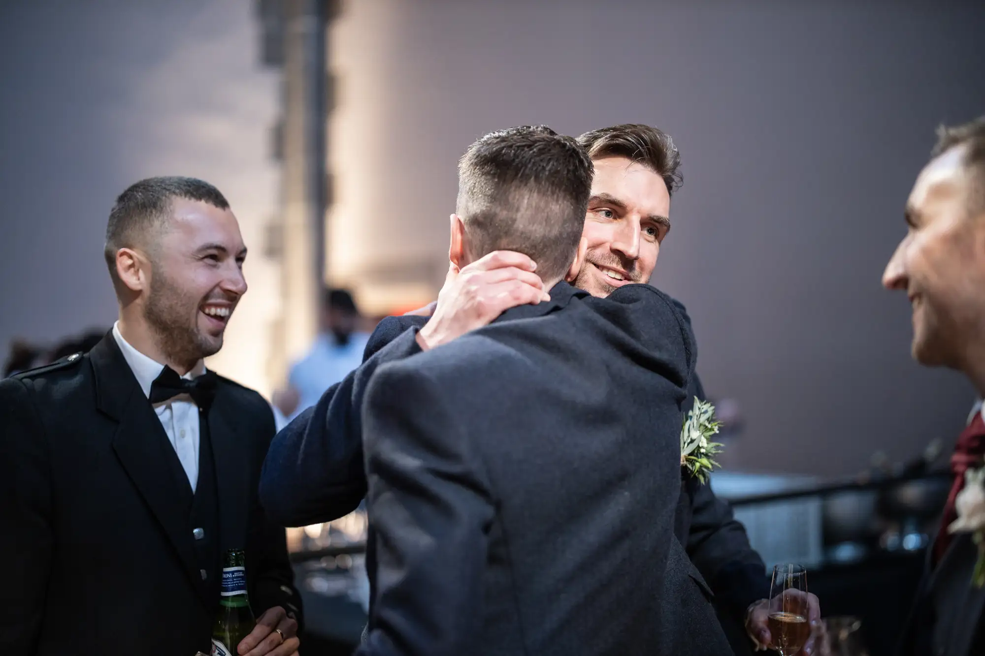Three men in formal attire at a social event, one embracing another while the third smiles, all engaged in a joyful conversation.