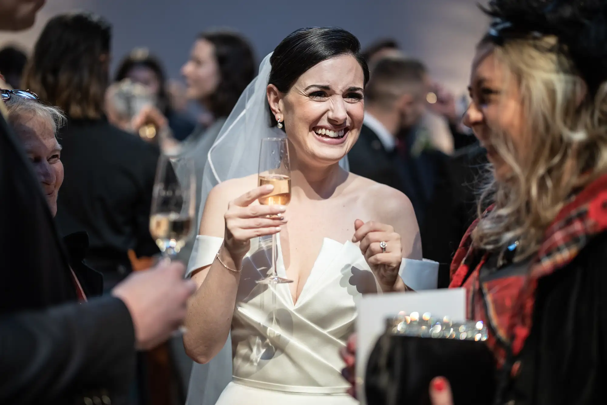 A bride in a white dress and veil laughs joyfully while holding a champagne glass, surrounded by guests at a wedding reception.