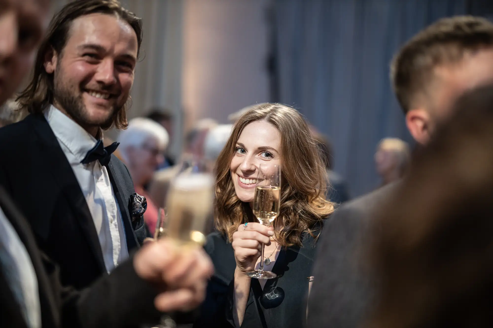 A woman smiling and holding a champagne glass at a social event, surrounded by people in formal attire.