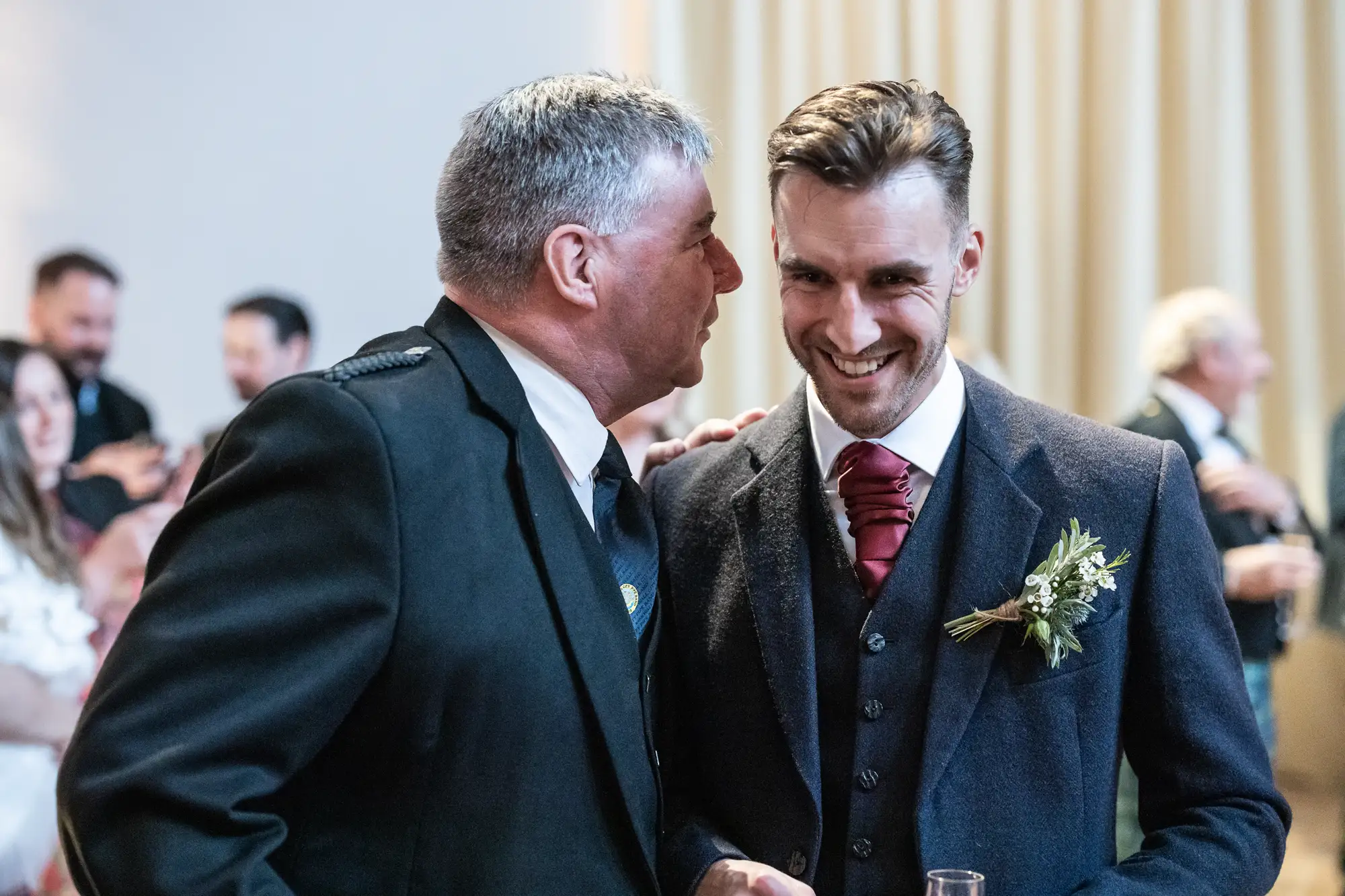 Two men smiling at each other at a wedding, one in a kilt and the other in a suit, both wearing boutonnieres.