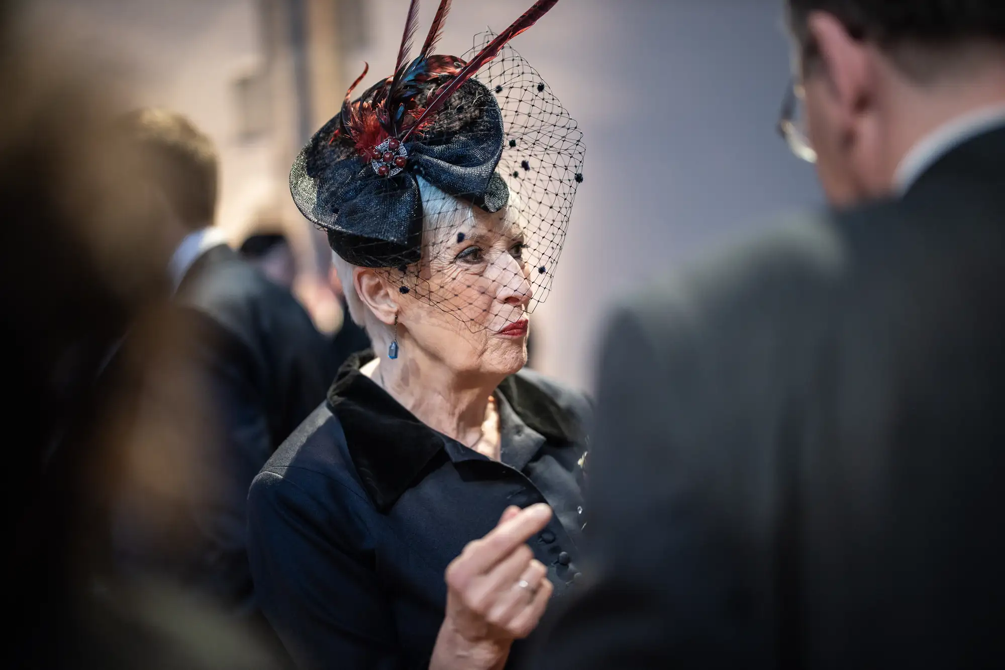Elderly woman in a stylish hat with a net, engaged in conversation at a social event.