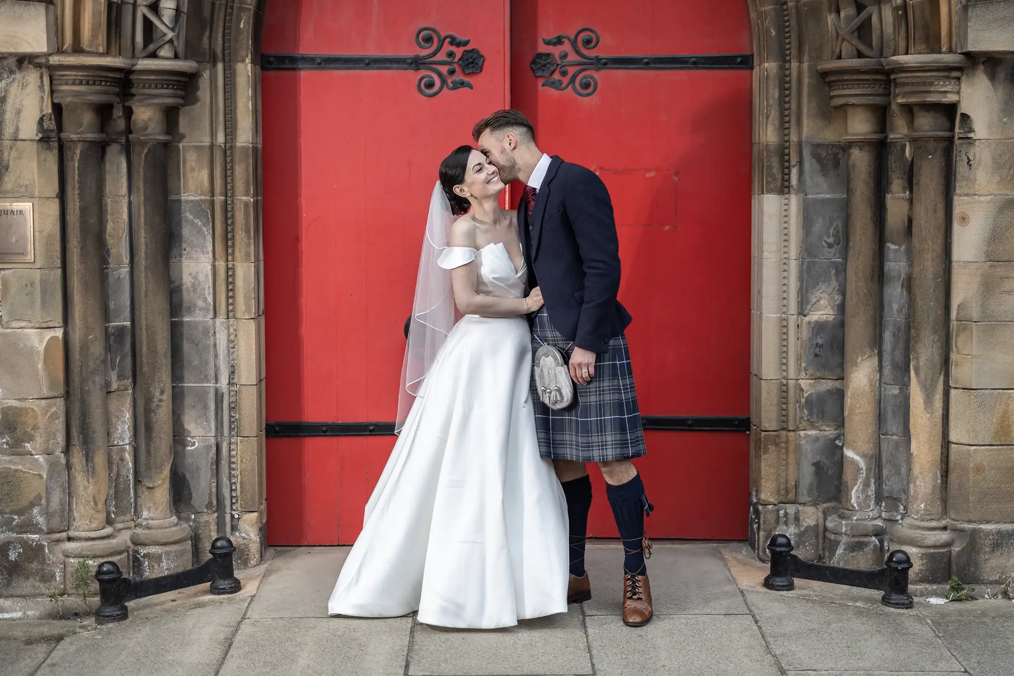A bride in a white gown and a groom in a kilt share a kiss in front of a historic building with a large red door.