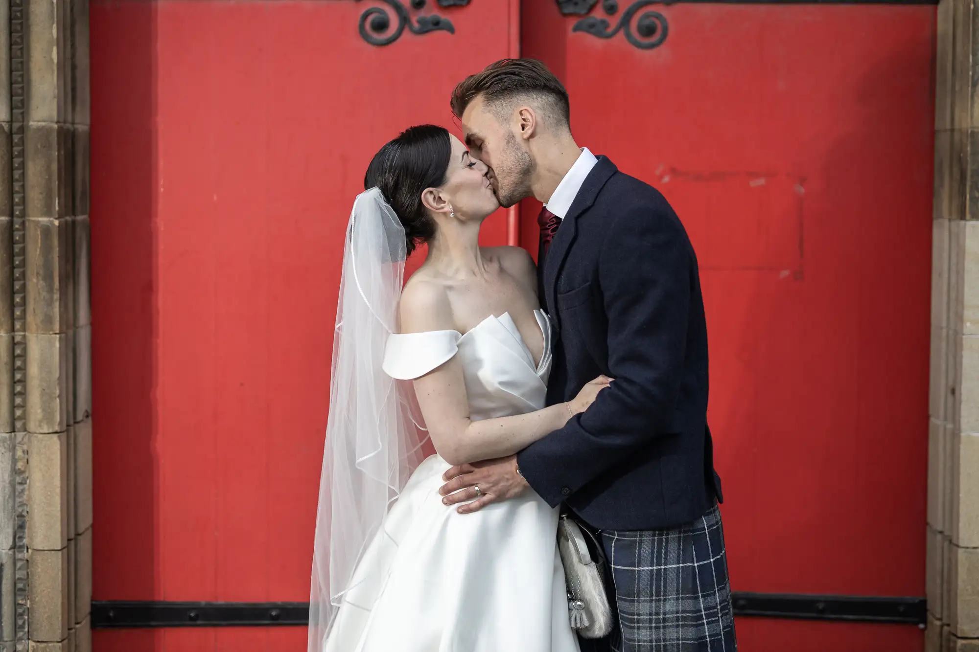 A newlywed couple kissing outside a red door, the bride in a white dress and veil, the groom in a kilt and jacket.