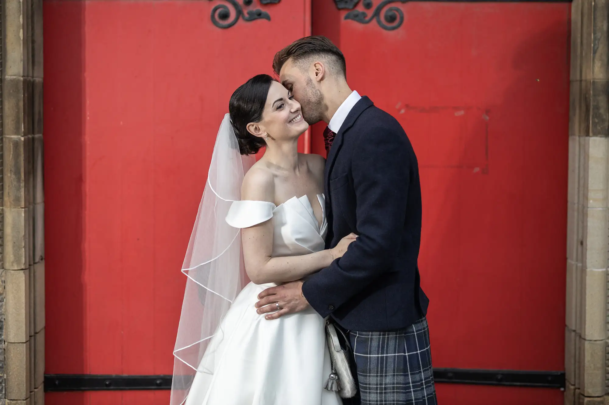 Bride and groom sharing a tender moment in front of a bright red door, with the groom wearing a kilt and the bride in a white dress and veil.