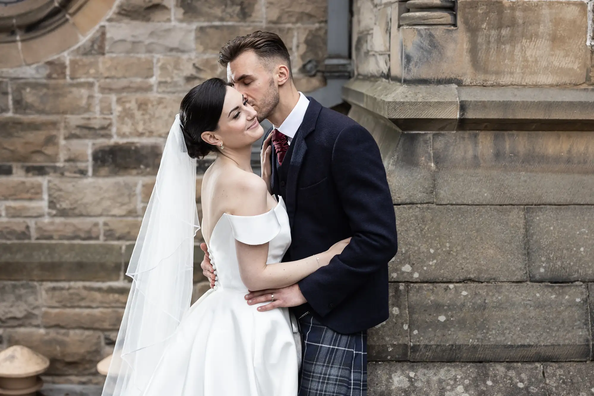 A bride in a white dress and a groom in a kilt embracing and kissing outside a stone building.