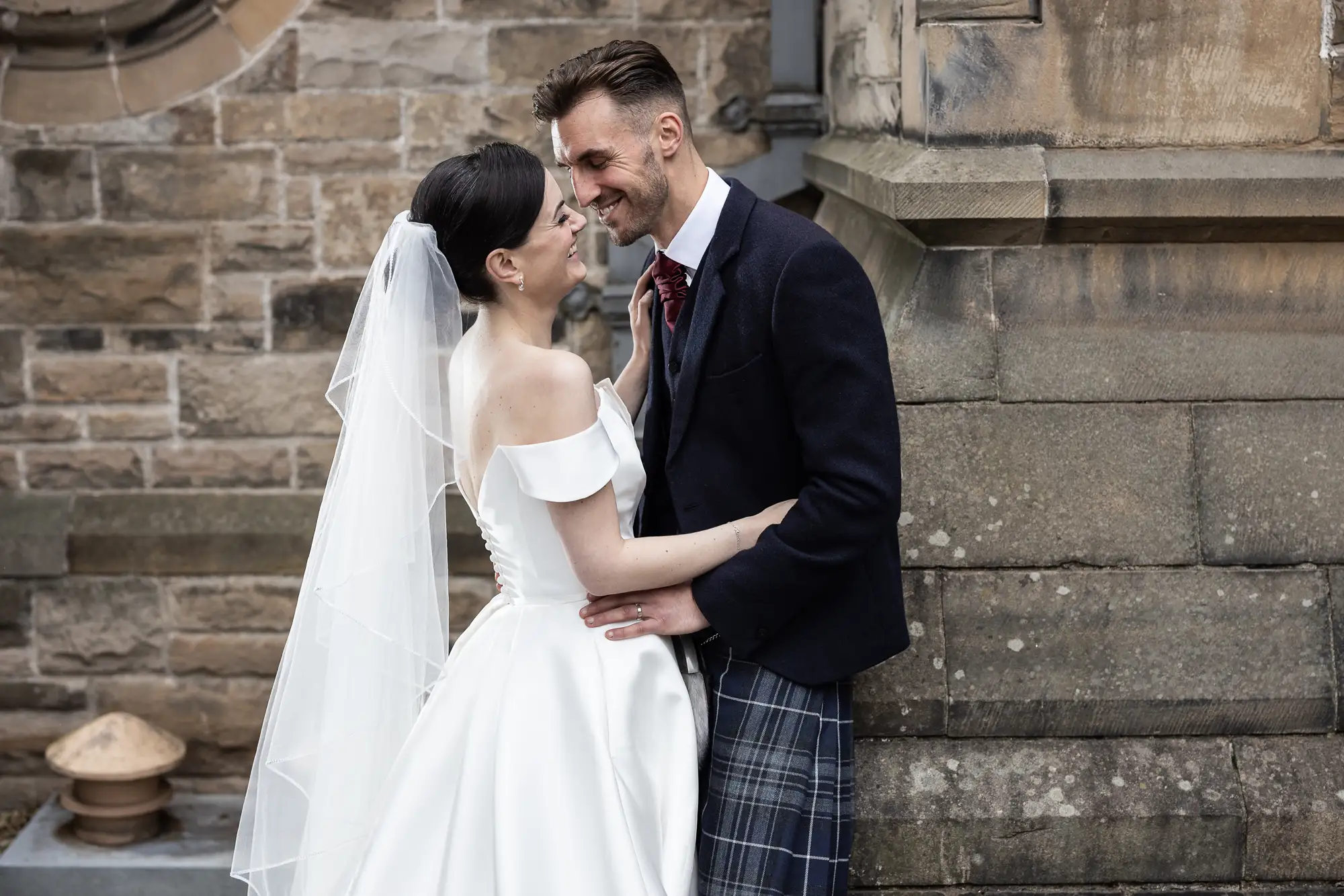 A bride and groom in wedding attire share a tender moment, with the groom in a kilt, against a historic stone building backdrop.