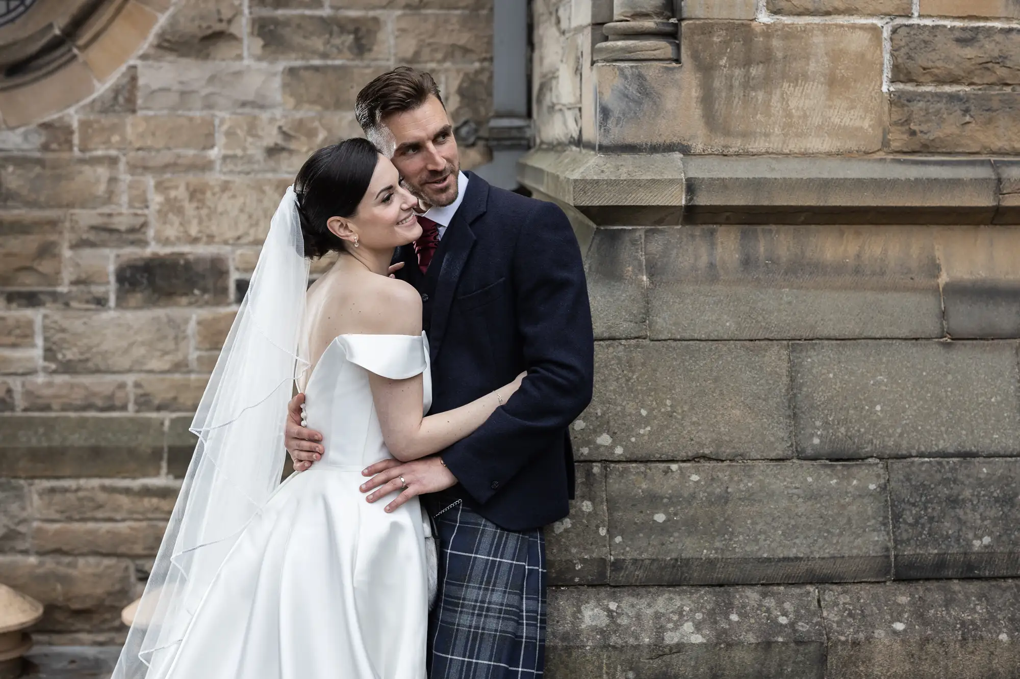A bride in a white dress and a groom in a tartan kilt embracing against a historic stone building background.