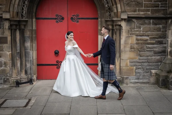 Vibrant wedding photos at Mansfield Traquair Edinburgh - Mansfield Traquair wedding photographer. Newlyweds Helen and James holding hands holding hands in front of a large red door, with the groom wearing a kilt and the bride in a white gown.