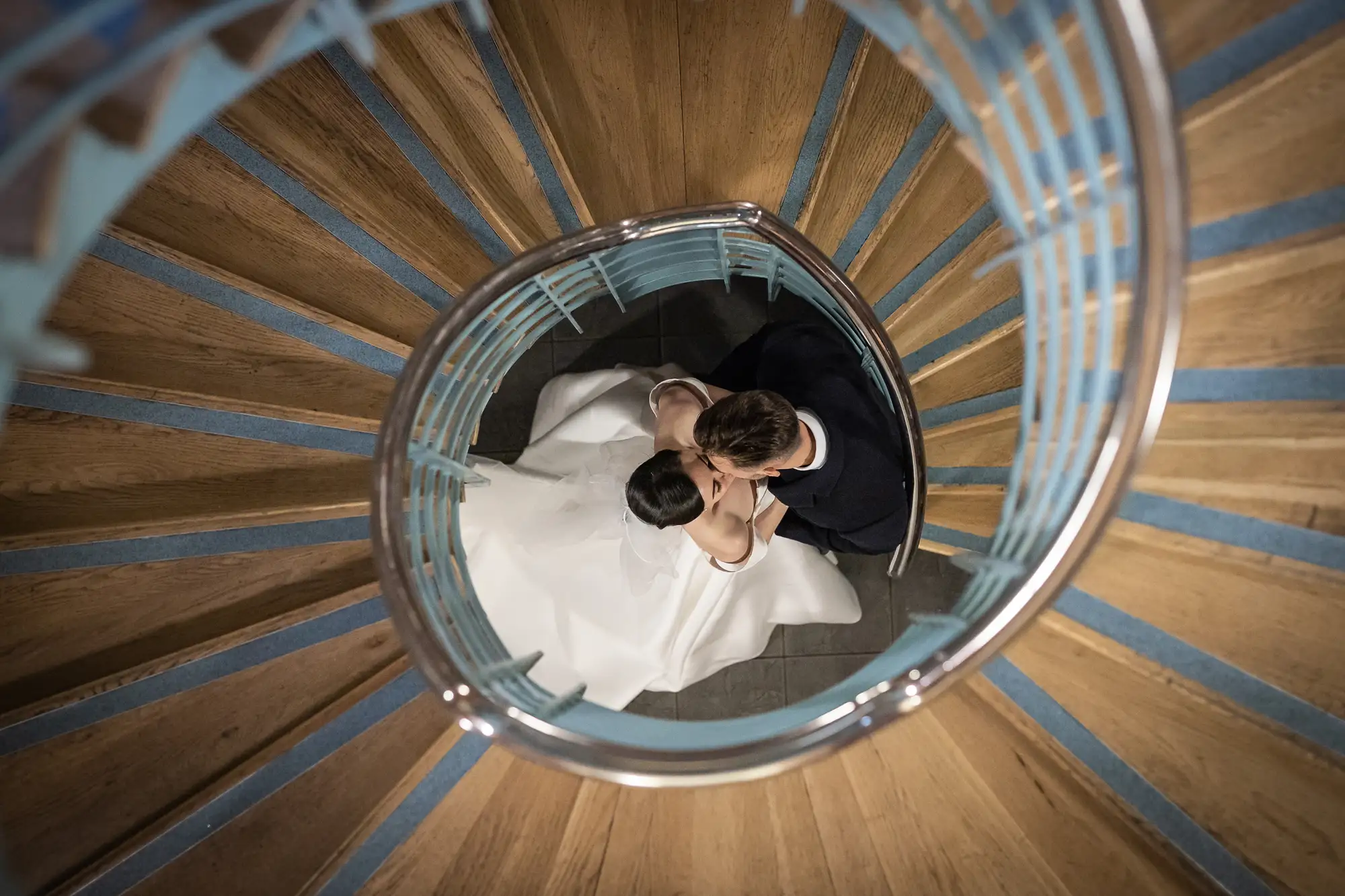 A bride and groom share a kiss at the bottom of a spiral staircase, viewed from above.