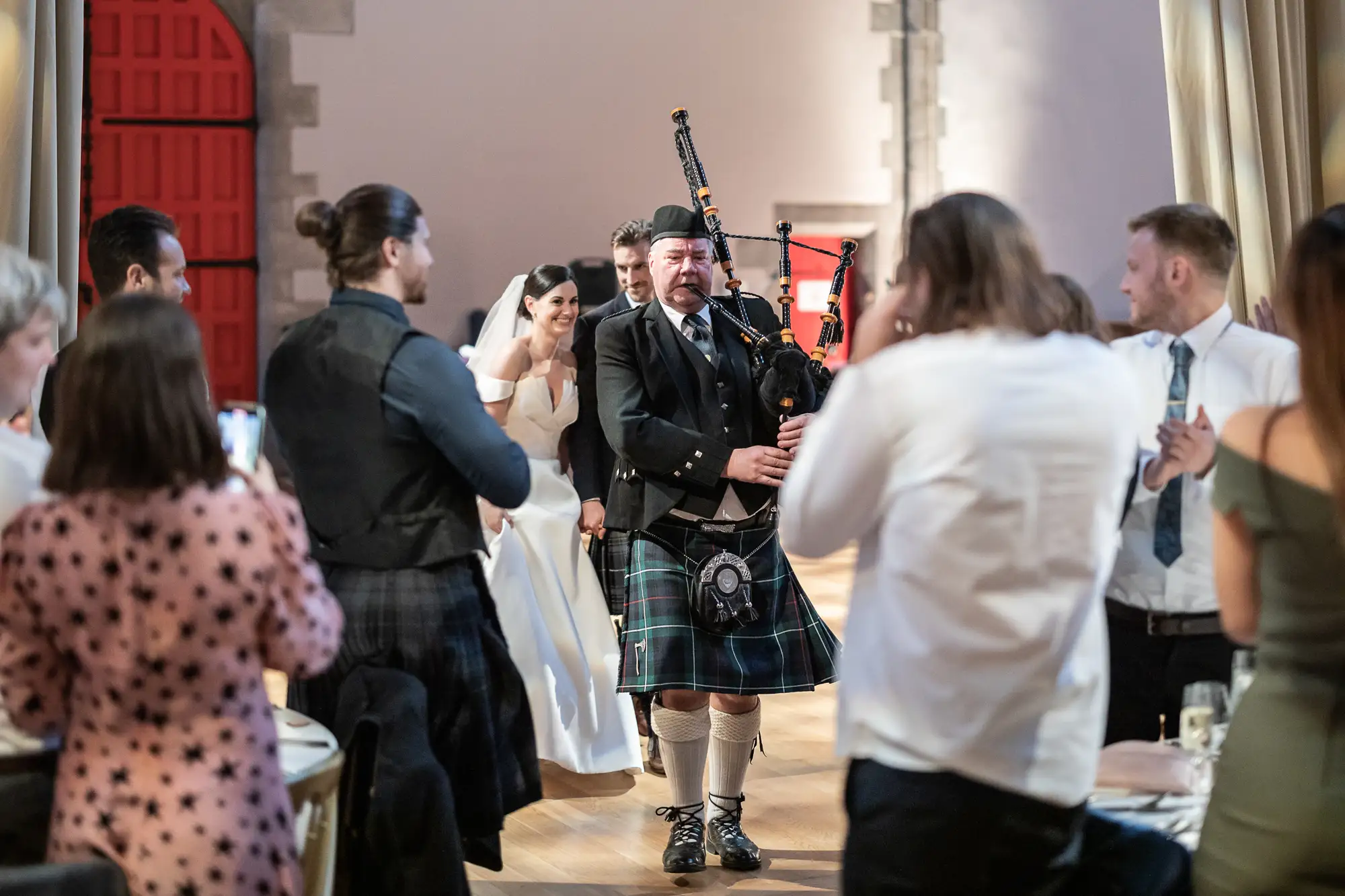 A bagpiper in traditional scottish attire performs at a wedding, with guests and a smiling bride and groom watching in the background.