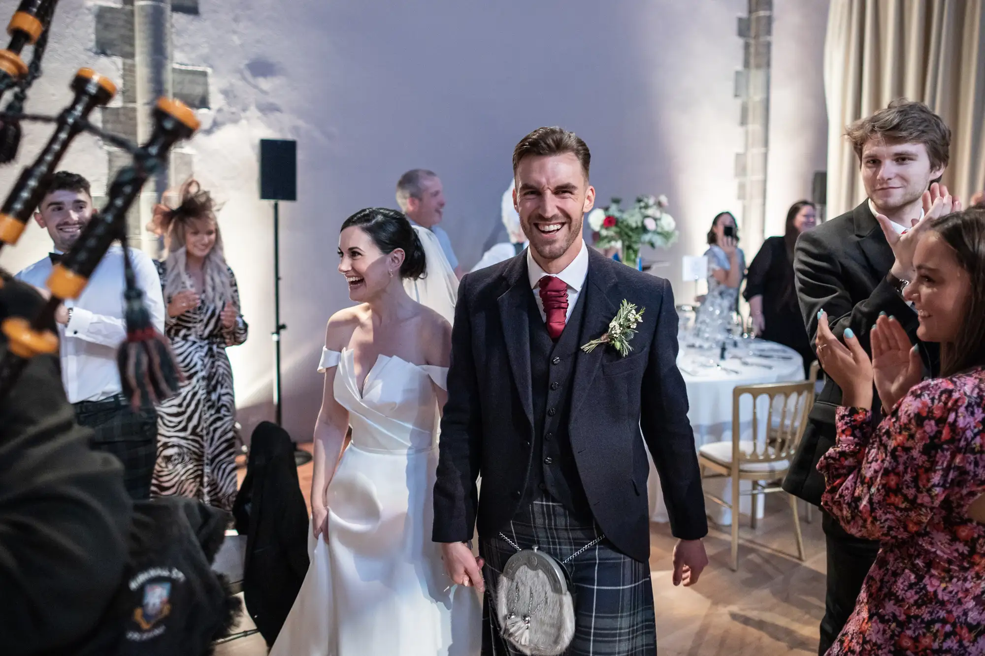A newlywed couple walking joyfully through a clapping crowd, the groom in a tartan kilt and the bride in a white dress.