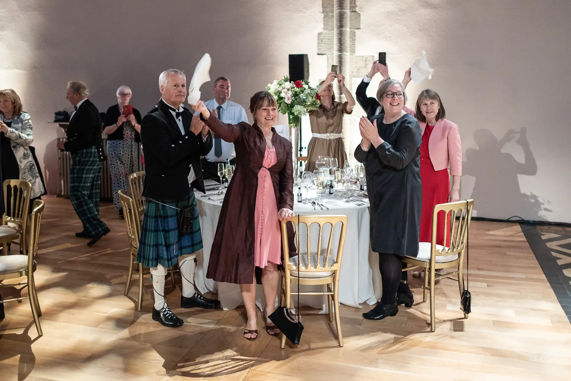 Guests dancing and clapping around a banquet table at a formal event, some wearing traditional scottish attire.