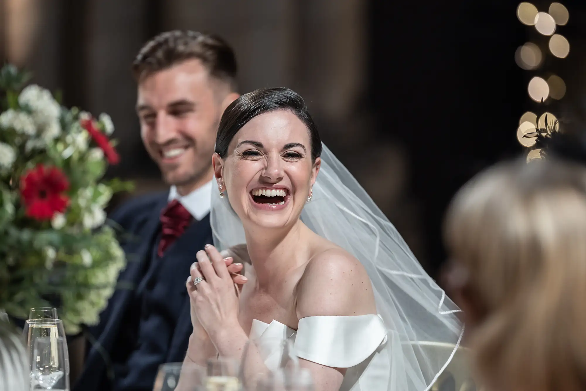 A bride in a white off-the-shoulder dress and veil laughing joyfully beside a smiling groom in a navy suit during a wedding reception.