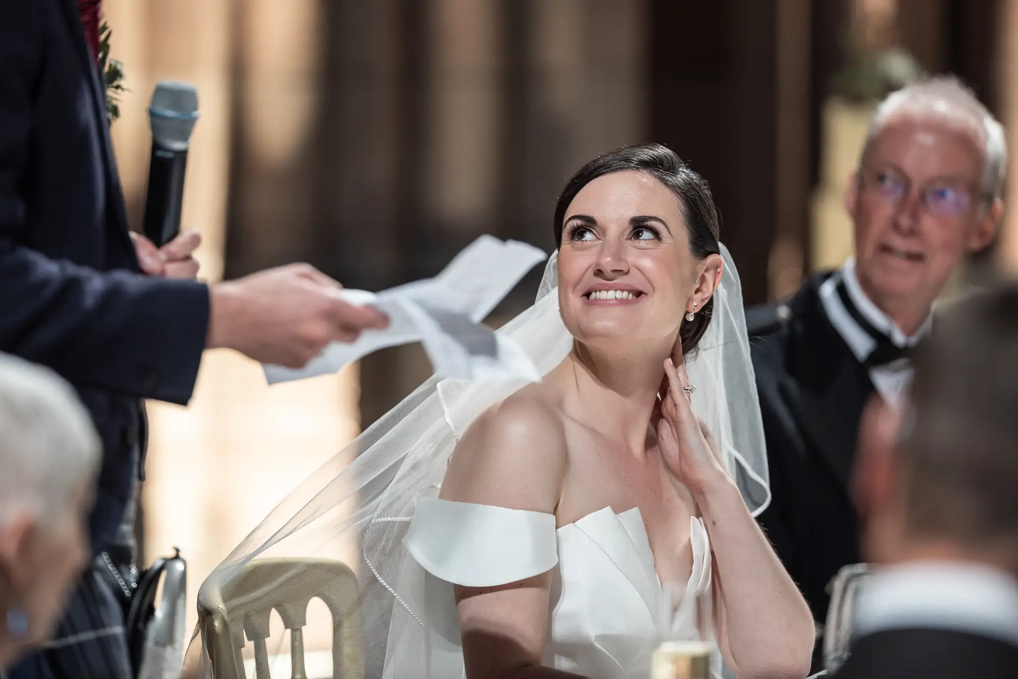 A bride, smiling joyfully, listens to a speech at her wedding reception, with guests around her and a speaker partially visible on the left.