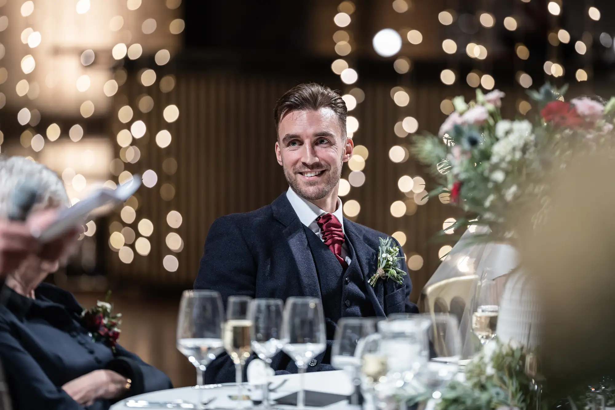 Man in a suit with a red tie and boutonniere smiling at a wedding reception, surrounded by guests and twinkling lights in the background.
