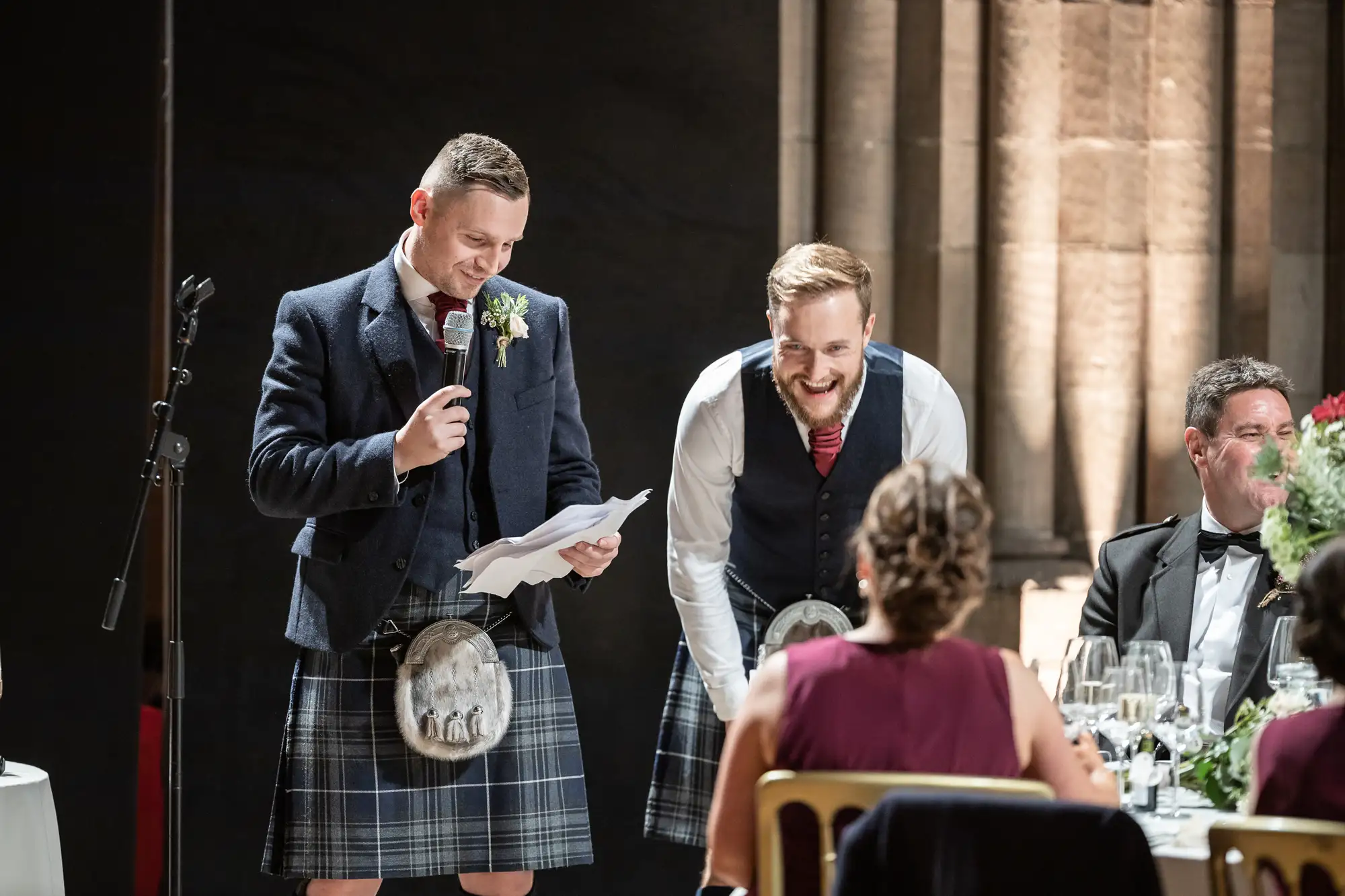 Two men at a wedding reception, one in a kilt reading a speech and smiling, the other listening and laughing, with guests seated around them.