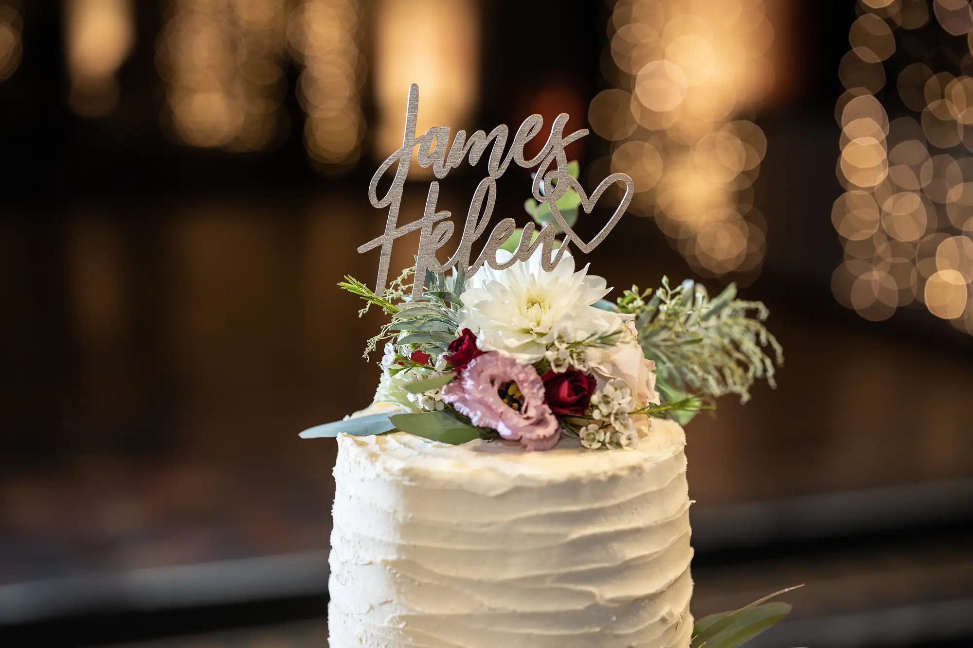 A wedding cake adorned with a "just married" topper and a mix of white and red flowers, set against a softly blurred background of golden lights.